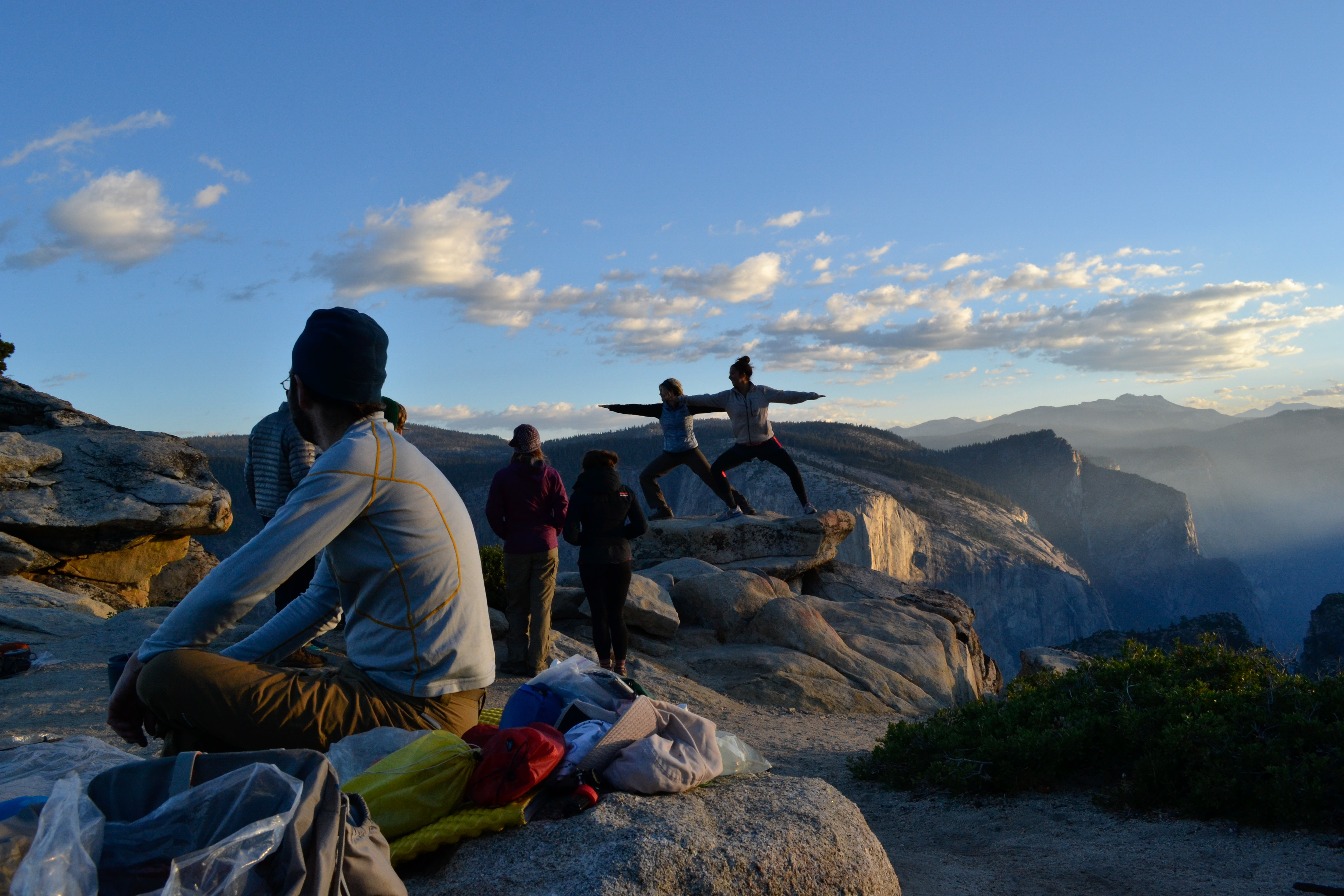 01 Jeannine Andre tmbtp image (1) A group of people - some seated, some standing - watch two women in a Warrior Yoga pose on a granite rock outcrop. There are large granite cliffs behind them, the skies have sparse clouds and sunlight streams in from the right hand corner.