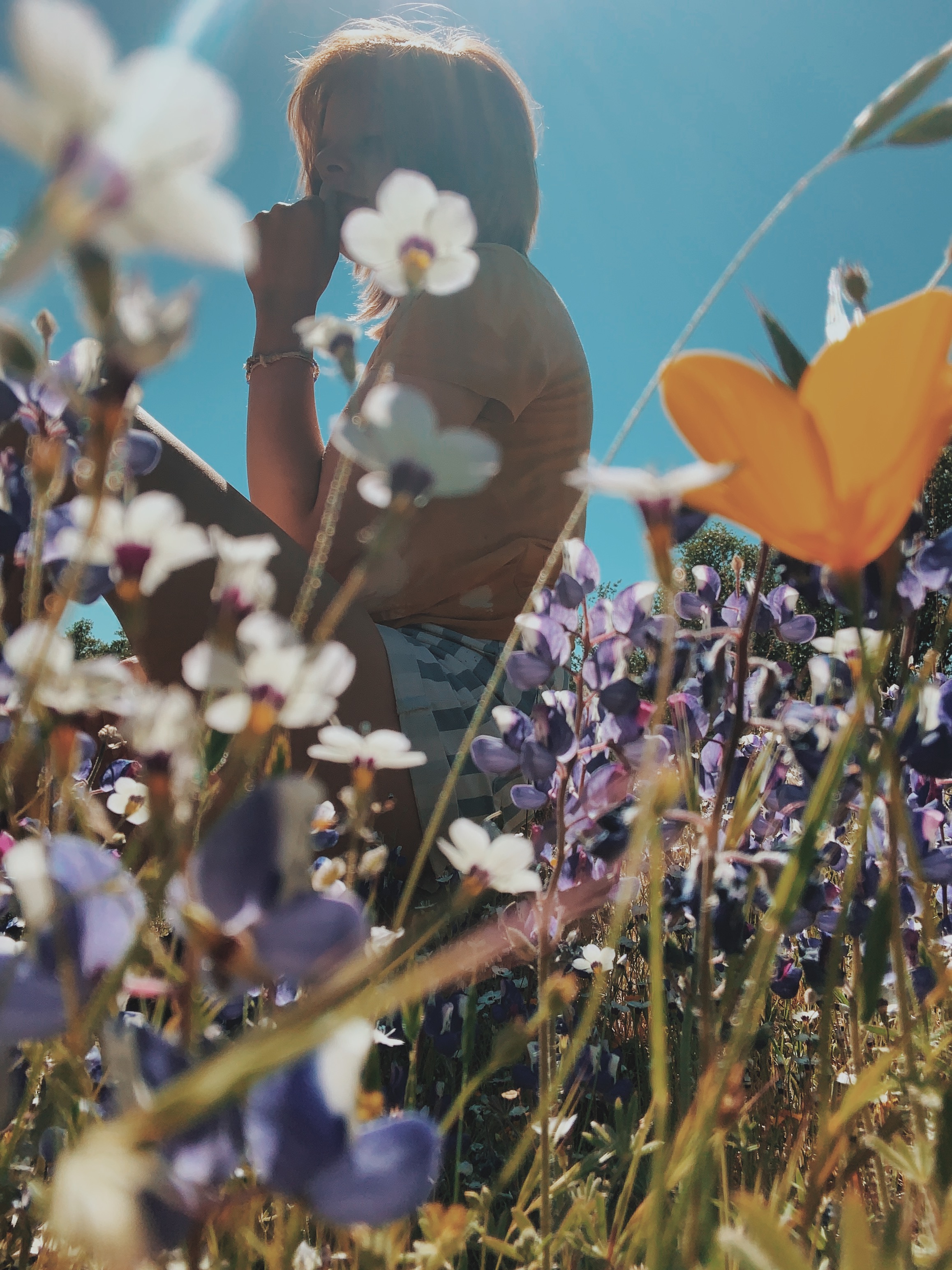 Processed with VSCO with c8 preset Close-up shot of wild flowers from their base. In the background, a young woman in an orange t-shirt and grey striped shorts is seated with her chin resting in her hand.