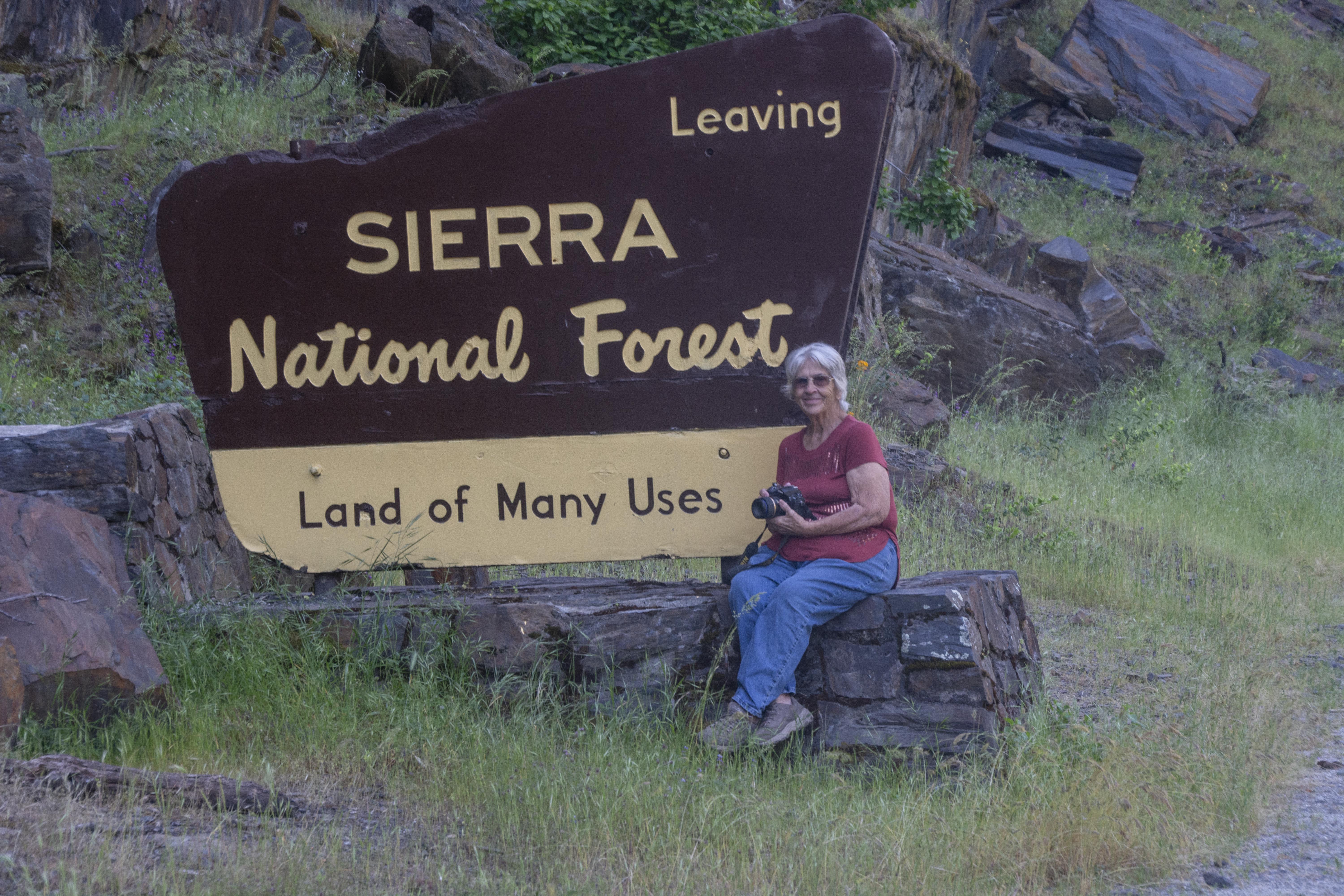 23 Jeanetta Philips tmbtp image Older woman is facing the camera, smiling and holding a camera while seated at the foot of sign reading "Leaving Sierra National Forest, Land of Many Uses".
