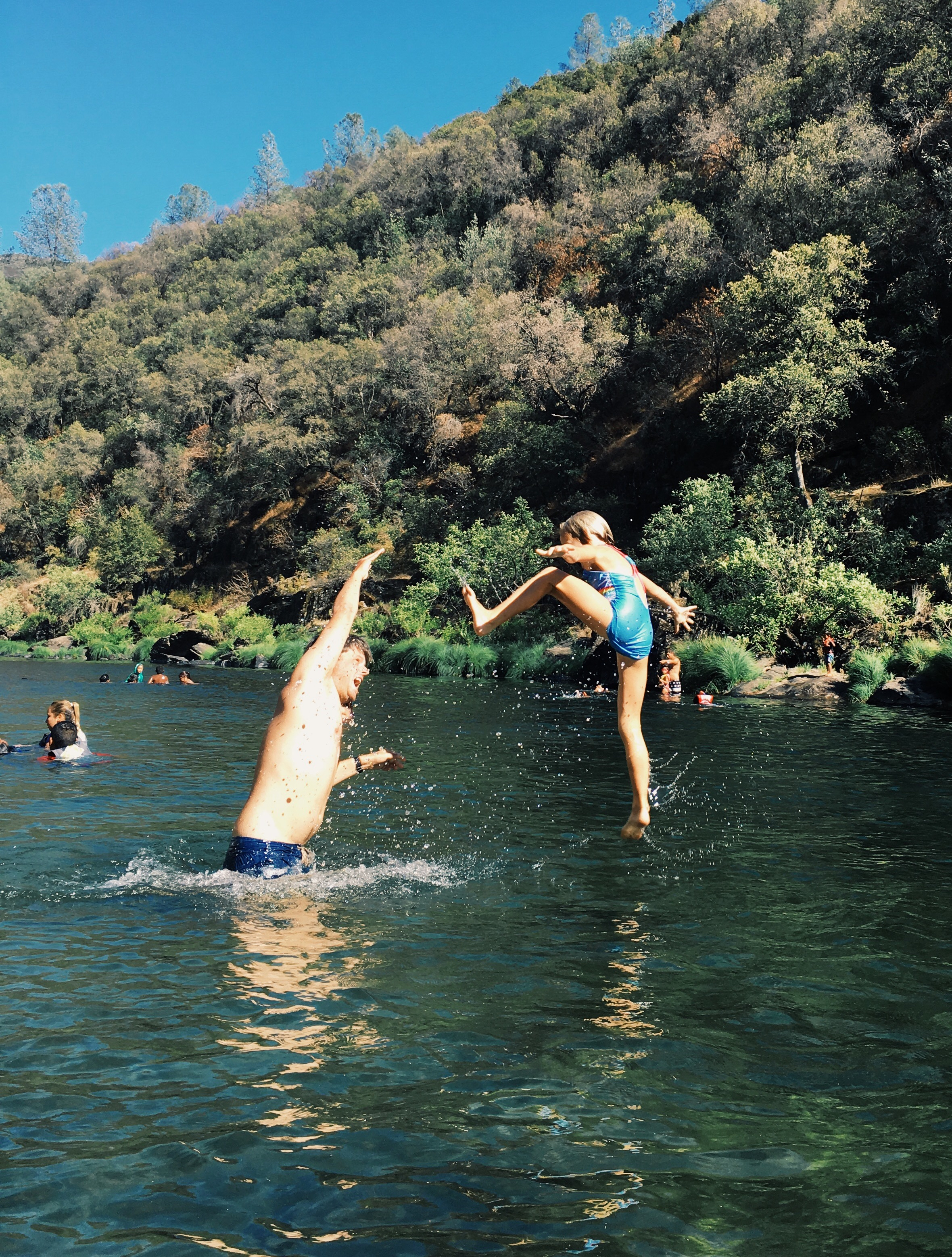 Processed with VSCO with g3 preset A young girl has her arms outreached as a man throws her upwards. They are in a river. Mountains and other swimmers can be seen in the background