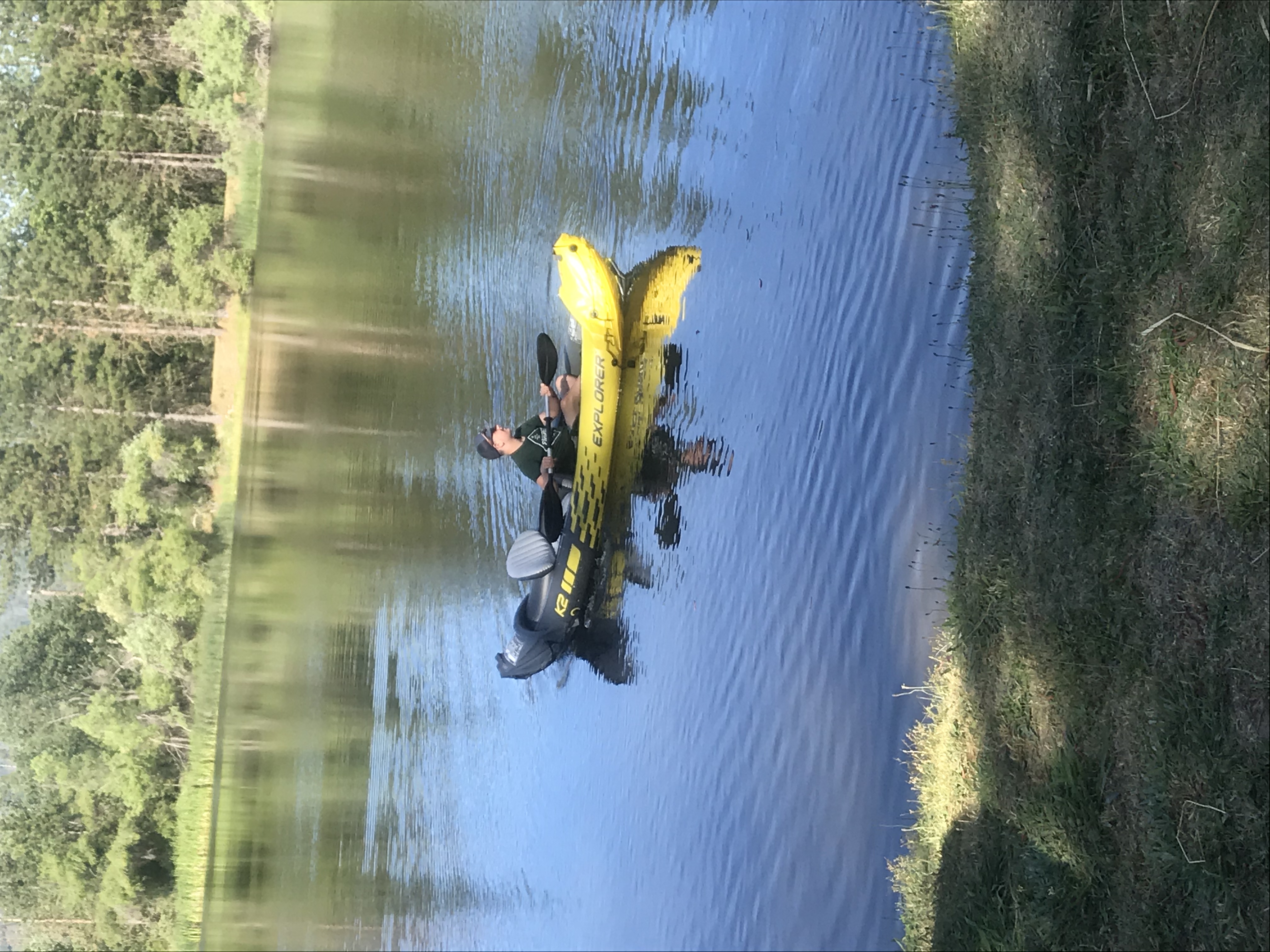 Tarah A man is seated in a yellow canoe in a small lake. He is looking out to the distance. Tall pine trees are in the background