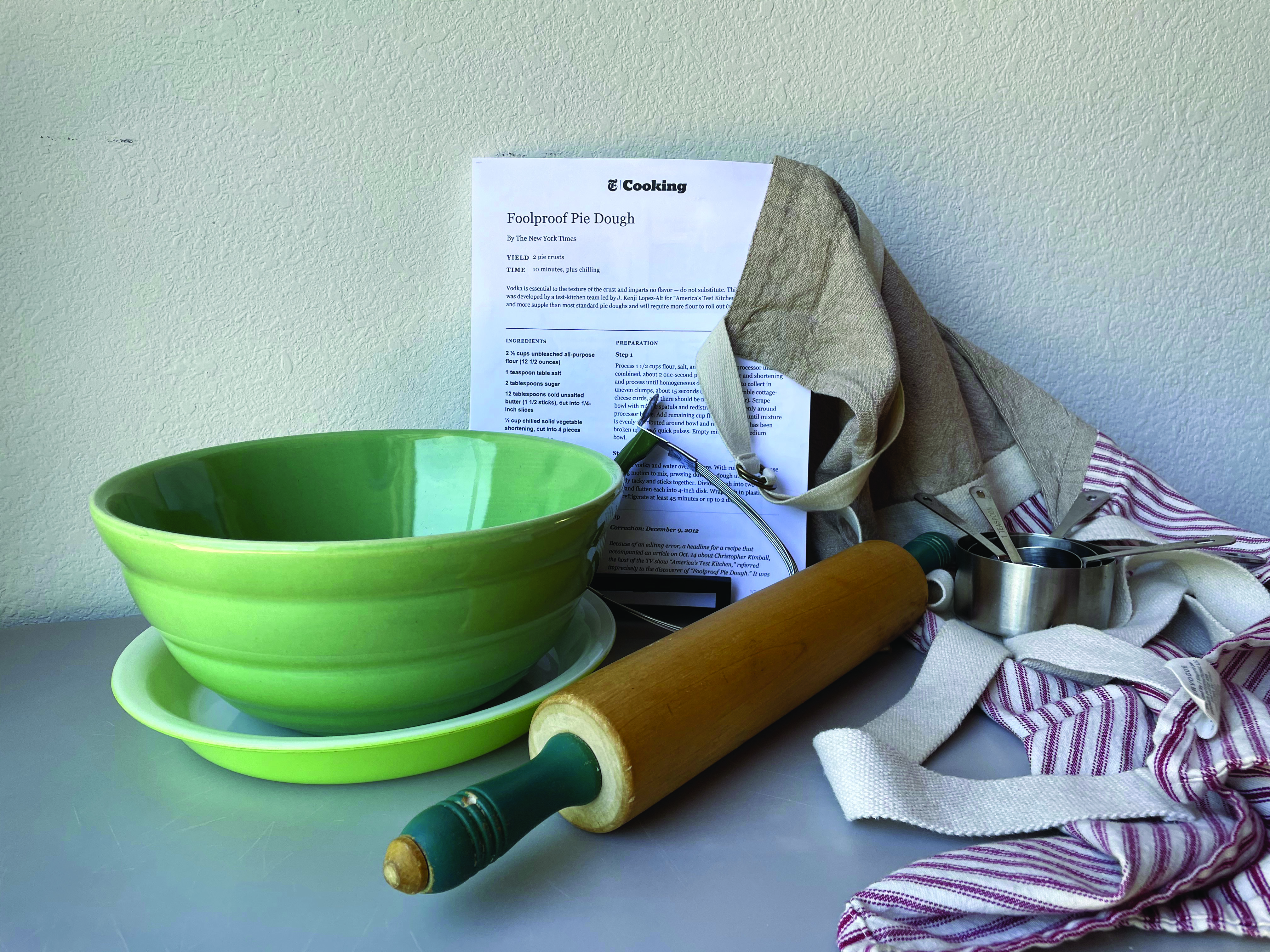 Photograph of a green ceramic mixing bowl, a recipe for Foolproof Pie Dough, a rolling pin and several aprons